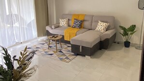 Ein modernes Wohnzimmer mit einem grauen Sofa, einem gelben Überwurf und gemusterten Kissen. Ein kleiner runder Tisch mit einer Fernbedienung steht auf dem Boden.