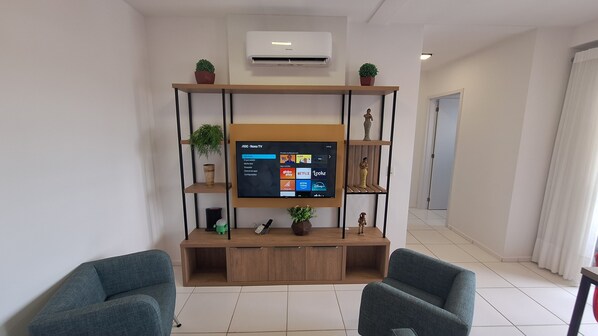 A living room with a flat-screen TV mounted on a wooden stand, two armchairs, a coffee table, and decorative items.