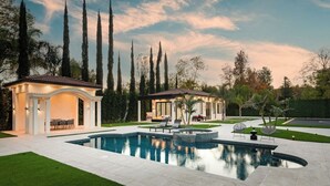 Un área de piscina exterior lujosa con un pabellón, una zona de estar cubierta y un césped cuidado.