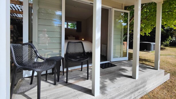 A covered patio with two chairs and a table, leading to a bedroom with a bed and a nightstand. - Happy Days and Relaxing Stays (Te Anau)