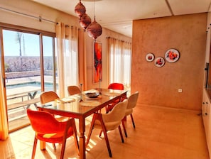 A dining area with a glass table, red and beige chairs, a decorative hanging light fixture, and a view of a pool and mountains through large windows. - Charming Villa Just 10 Minutes from Essaouira – Peace, Pool & Privacy (Essaouira)