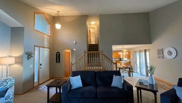 A spacious living room with a dark-colored sofa, a coffee table, and a dining area with a small table and chairs. A staircase leads to an upper level with a kitchen visible in the background.
