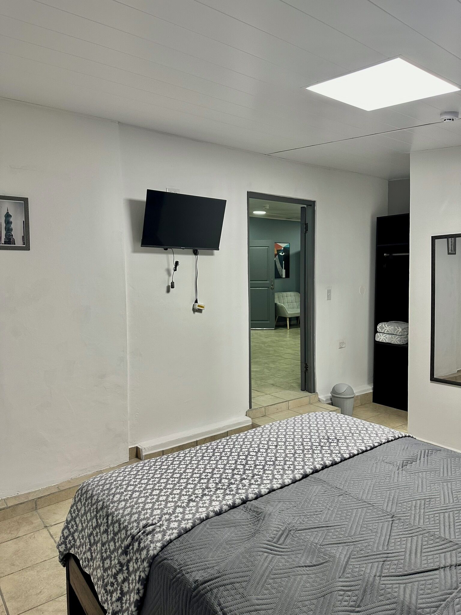 Een slaapkamer met een bed, een flatscreen televisie aan de muur, een deur naar een badkamer en een kleine prullenbak.