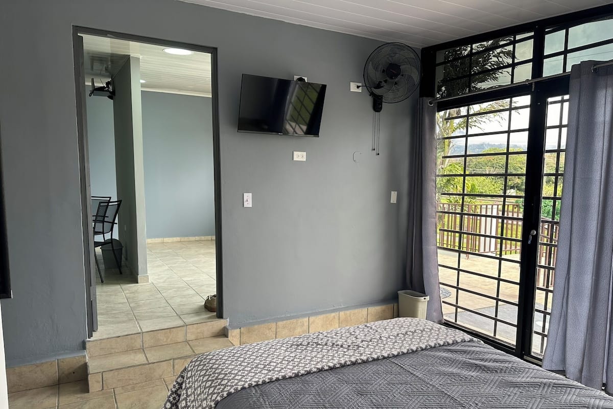 A bedroom with a bed, a TV, a fan, and a window with a view of the outdoors.