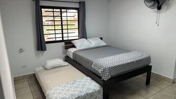 Una habitación pequeña y limpia con cama, mesita de noche, ventana con cortinas y un ventilador de pared.