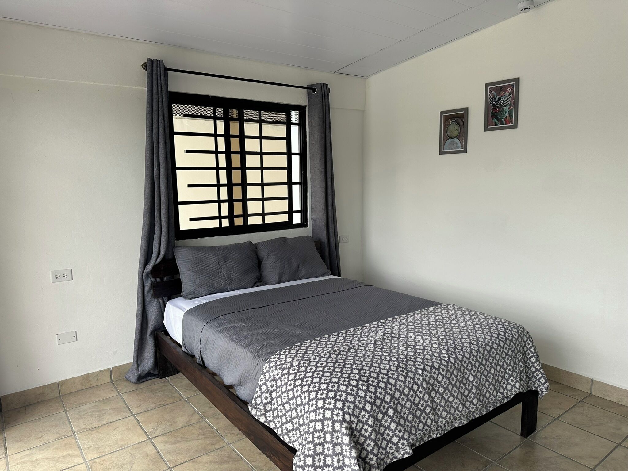 Ein Einzelbett mit gemustertem Überwurf, ein Fenster mit Gitterstruktur und zwei gerahmte Bilder an der Wand.
