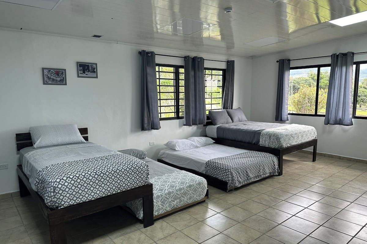 A room with three beds, large windows with curtains, and a tiled floor.