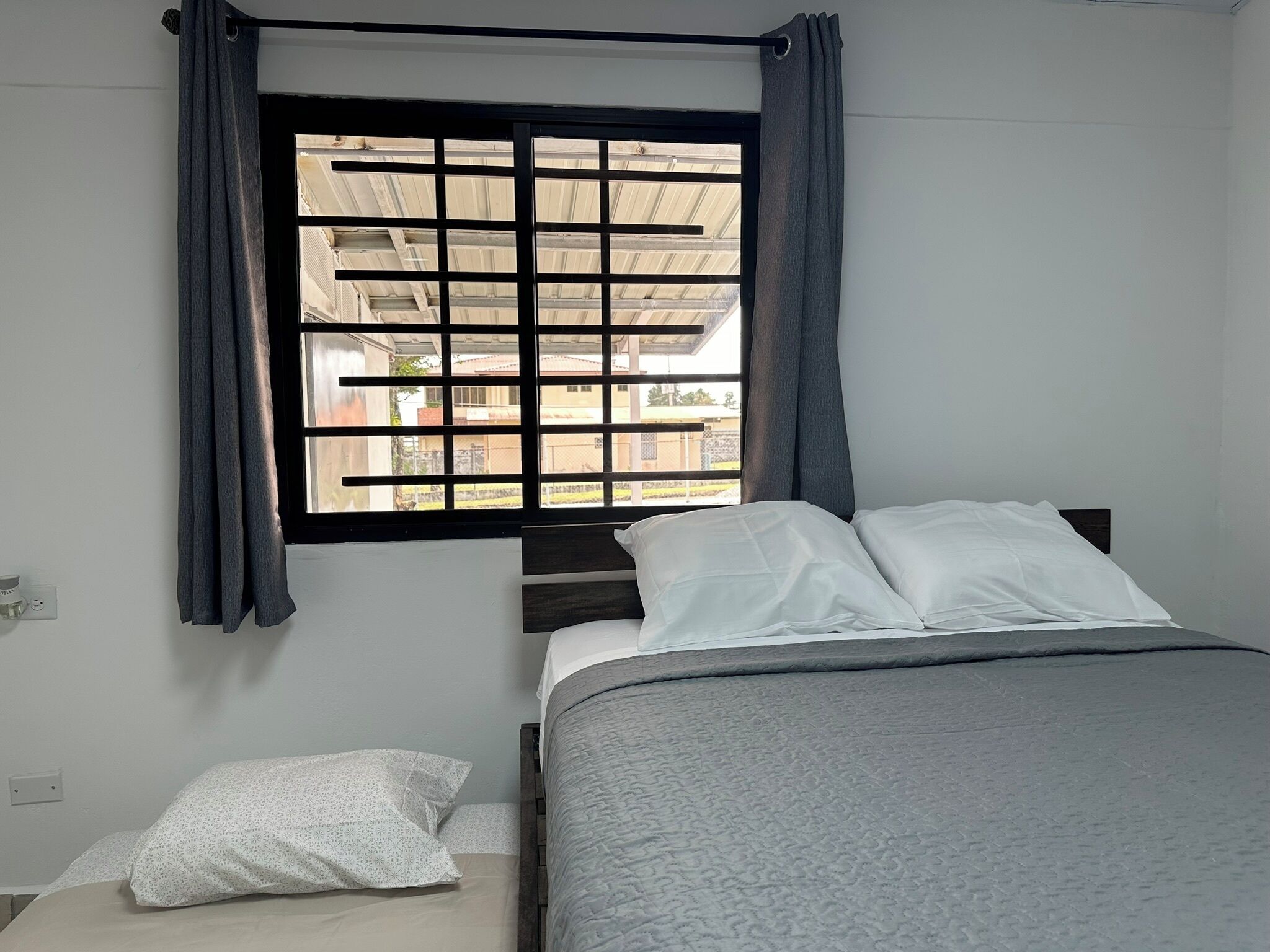 Una camera da letto con un letto, una finestra che offre vista su edifici e una presa di corrente.
