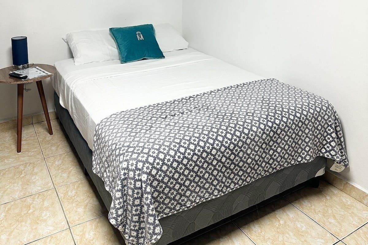 A single bed with a patterned bedspread, a teal pillow, and a bedside lamp on a small wooden table.