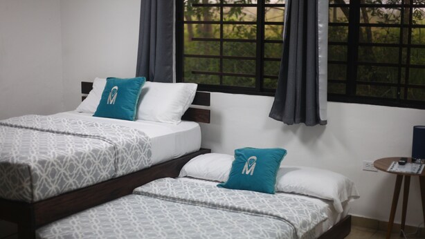 Habitación de hotel con dos camas, cada una con ropa de cama estampada y almohadas color turquesa. Hay una mesita con un control remoto y un enchufe.