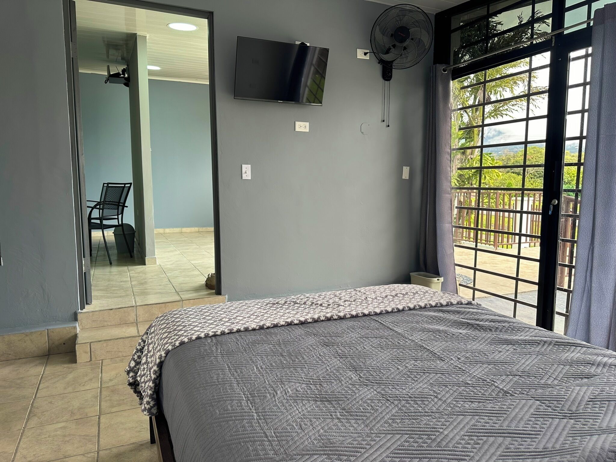Ein Schlafzimmer mit Bett, Wandfernseher, Standventilator und Blick ins Freie durch ein Fenster.