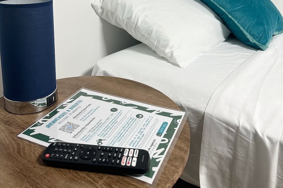 A hotel room with a bed, a blue lamp, a remote control, and a welcome brochure on a wooden table.