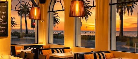 Un restaurante con mesas y sillas dispuestas para comer, amplias ventanas con vistas a la puesta de sol y palmeras, y lámparas colgantes.