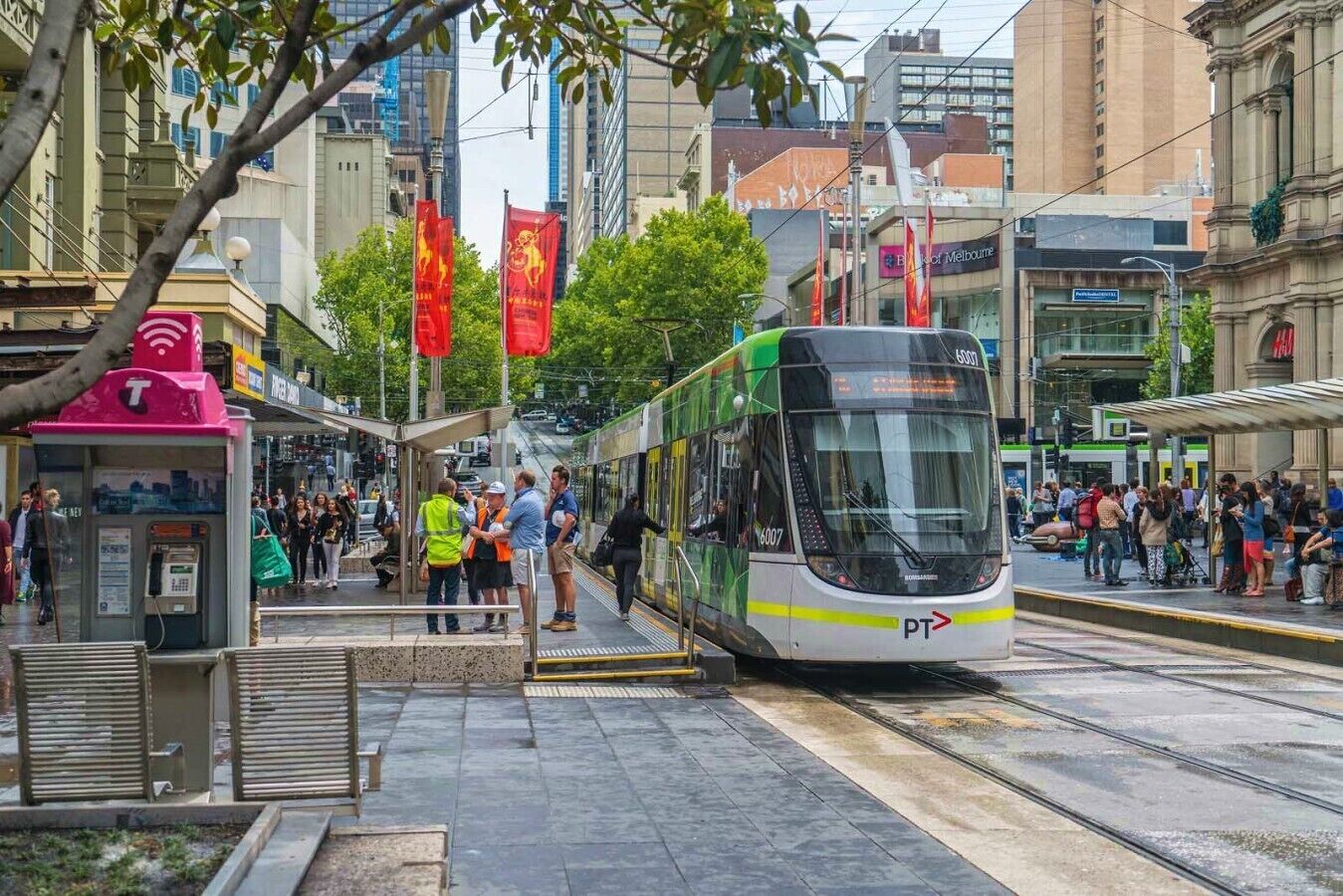 Een tramhalte met een moderne tram, wachtende passagiers en stedelijke gebouwen op de achtergrond.
