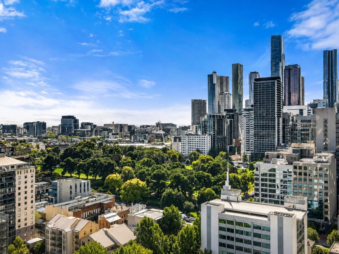 A cityscape with modern high-rise buildings, a park area with trees, and a clear blue sky.