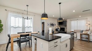 Une cuisine moderne dotée d’un îlot central, de luminaires suspendus noirs et d’un coin repas avec une table et des chaises en bois.
