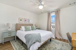 A bedroom with a bed, bedside tables, a ceiling fan, a window with curtains, and framed pictures on the wall.