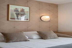 A hotel room with a bed, pillows, a framed picture on the wall, and a wall-mounted light fixture.
