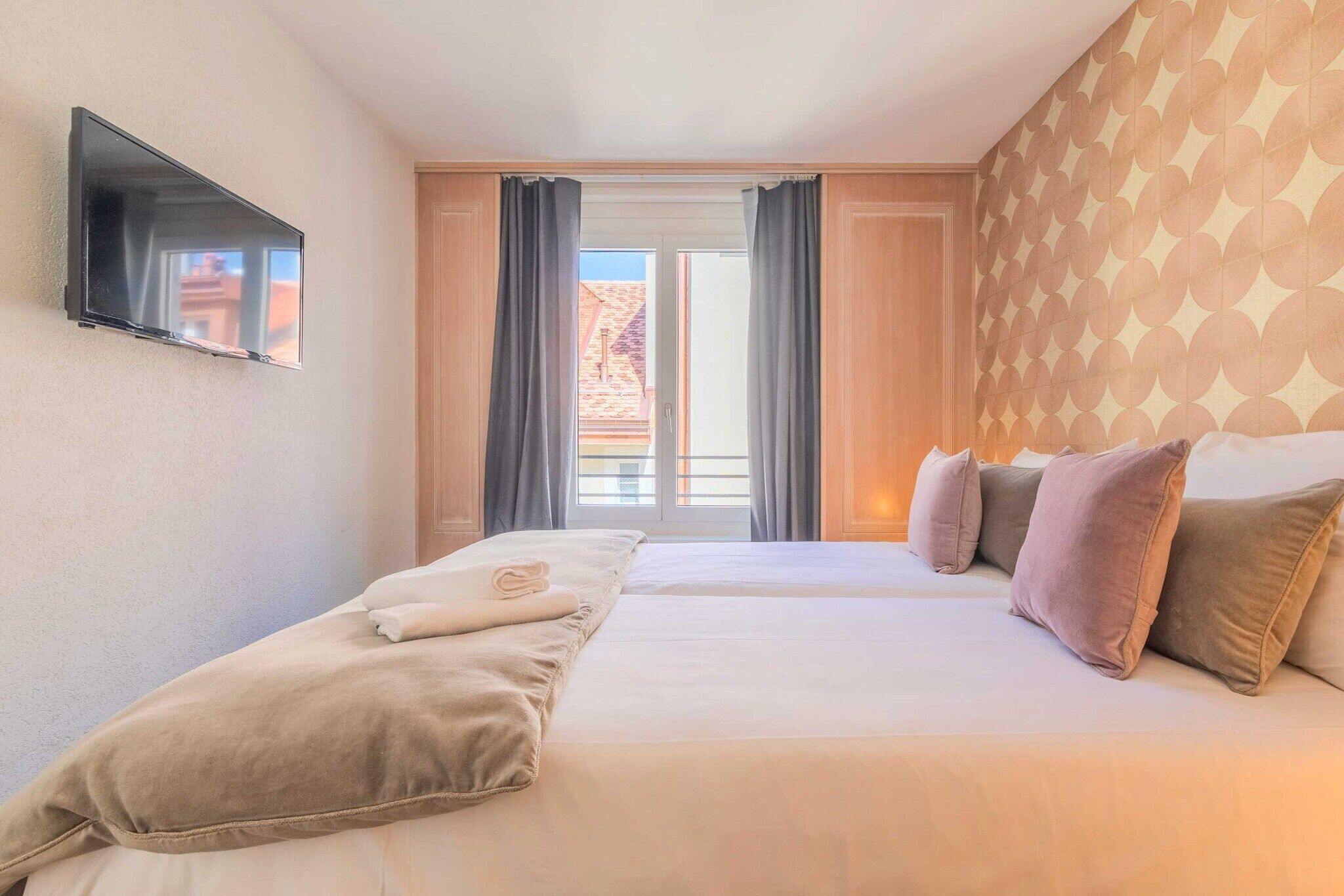 A hotel room with two beds, a flat-screen TV mounted on the wall, and a view of the outside through a large window with curtains.