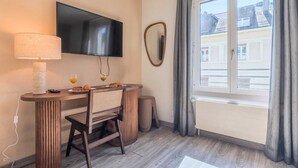 Une chambre d’hôtel avec un téléviseur fixé au mur, une table à manger avec une chaise, une lampe et une fenêtre avec des rideaux.