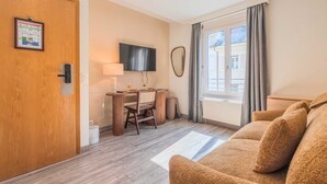 Une chambre d’hôtel avec une porte en bois, un bureau avec une lampe, une télévision, une grande fenêtre avec des rideaux et un canapé.
