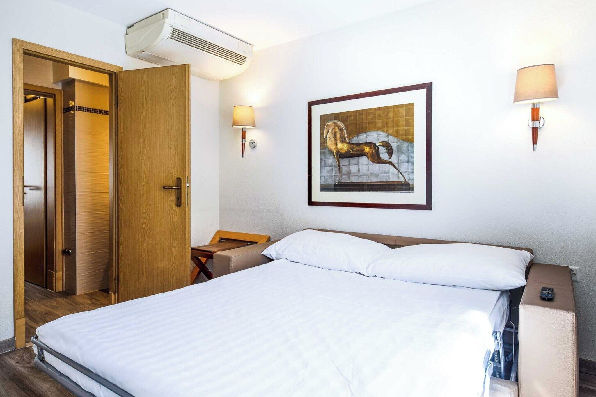 Een hotelkamer met een bed, een houten deur, een wandairconditioner, een lijst met een afbeelding en twee wandlampen.