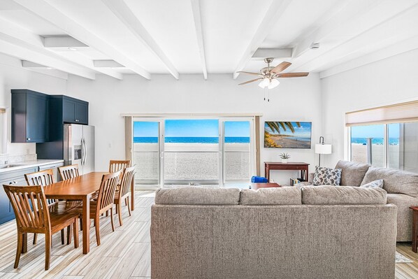 A spacious living area with a large window overlooking a beach, a ceiling fan, and a dining area with wooden furniture.