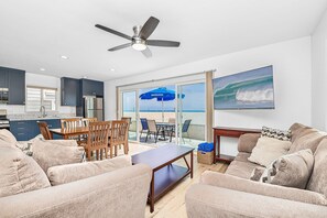A living room with a ceiling fan, a kitchen with dark cabinets, a dining area, and a large sliding glass door leading to a balcony with a beach view.