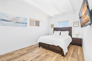 A bedroom with a wooden floor, a bed with white bedding, two windows, and artwork on the walls.