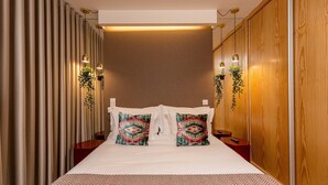 Hotellihuone, jossa on sänky, kaksi koristeellista tyynyä, yöpöytä, seinälle kiinnitetty kasvi ja puupanelilla verhoiltu seinä.