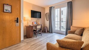 A hotel room with a wooden door, a desk with a computer, a TV, a sofa, and a large window with curtains. - Appartement moderne - AC-1BR-4P- Genève (Genève)