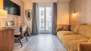 A hotel room with a flat-screen TV, a wooden desk with a laptop, a chair, a sofa, a window with curtains, and a patterned wall. - Appartement moderne - AC-1BR-4P- Genève (Genève)