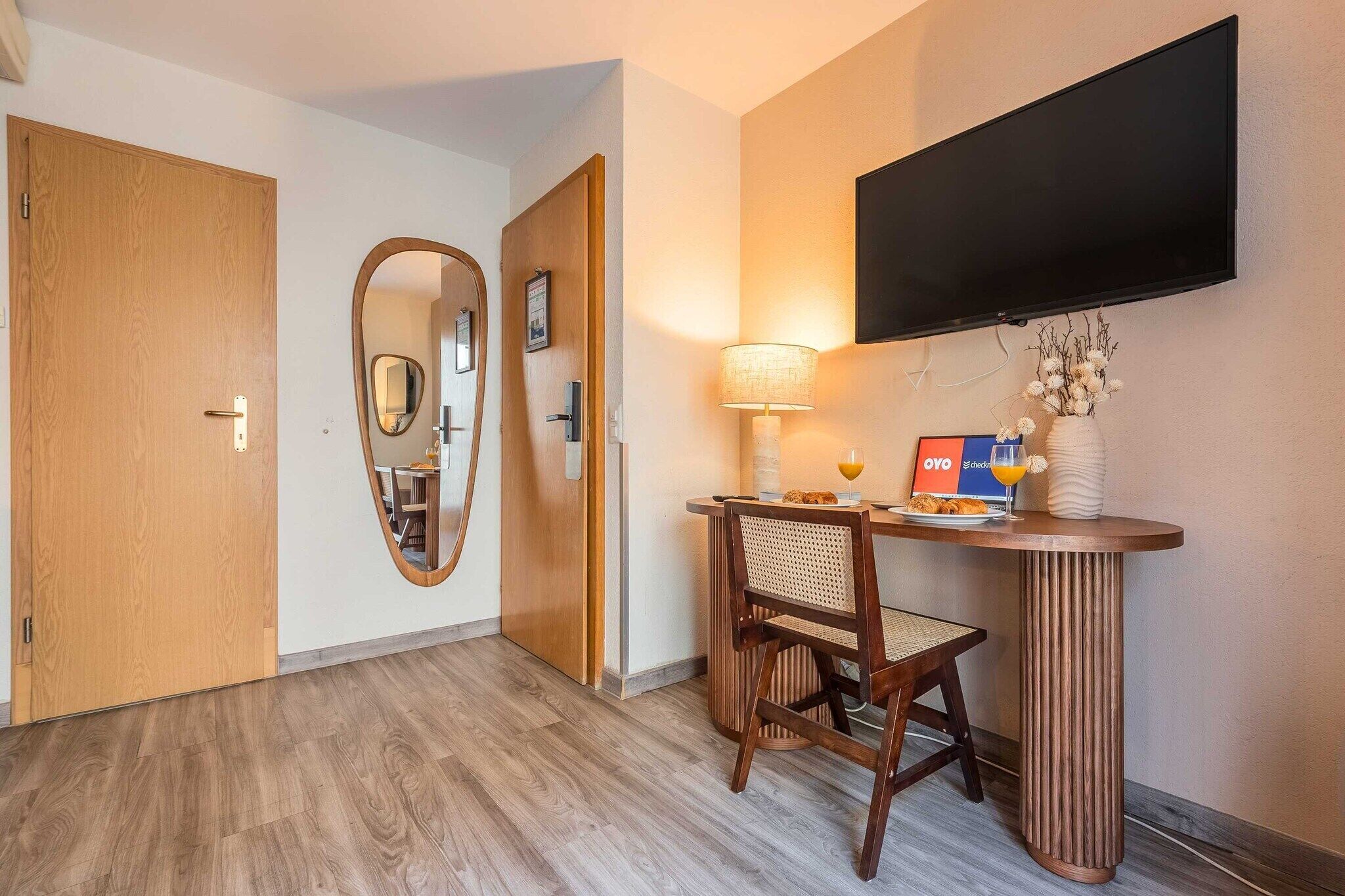 Una camera d'albergo con una porta in legno, uno specchio, una scrivania con lampada, una sedia e una TV a schermo piatto montata a parete.