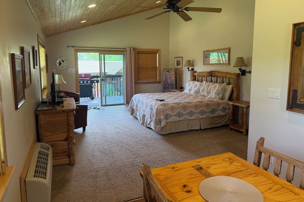 A bedroom with a bed, a ceiling fan, a desk with a lamp, a heater, and a sliding glass door leading to an outdoor area.