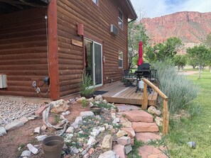A wooden cabin with a deck, a barbecue grill, and a mountain backdrop.