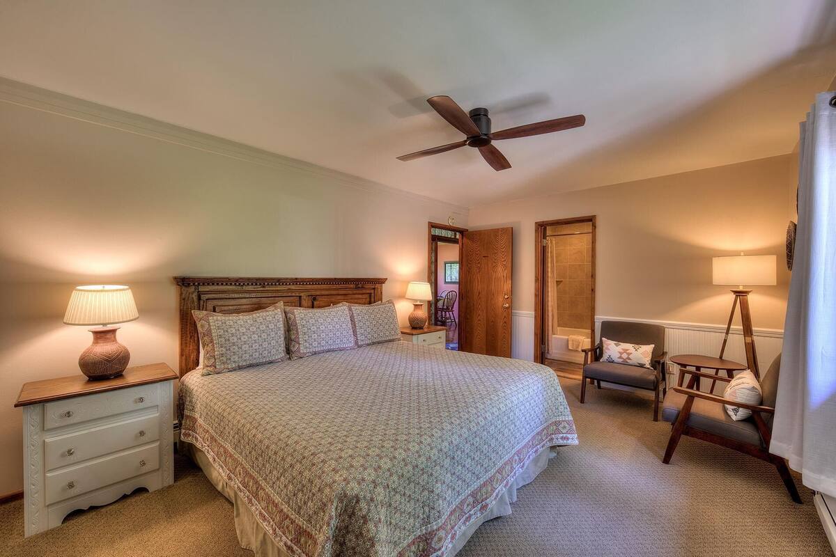 A bedroom with a bed, bedside lamps, a ceiling fan, a chair, and a door leading to another room.