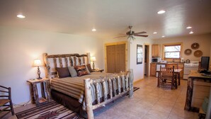 A bedroom with a bed, bedside tables, a wooden headboard, a ceiling fan, and a view into the adjacent kitchen.