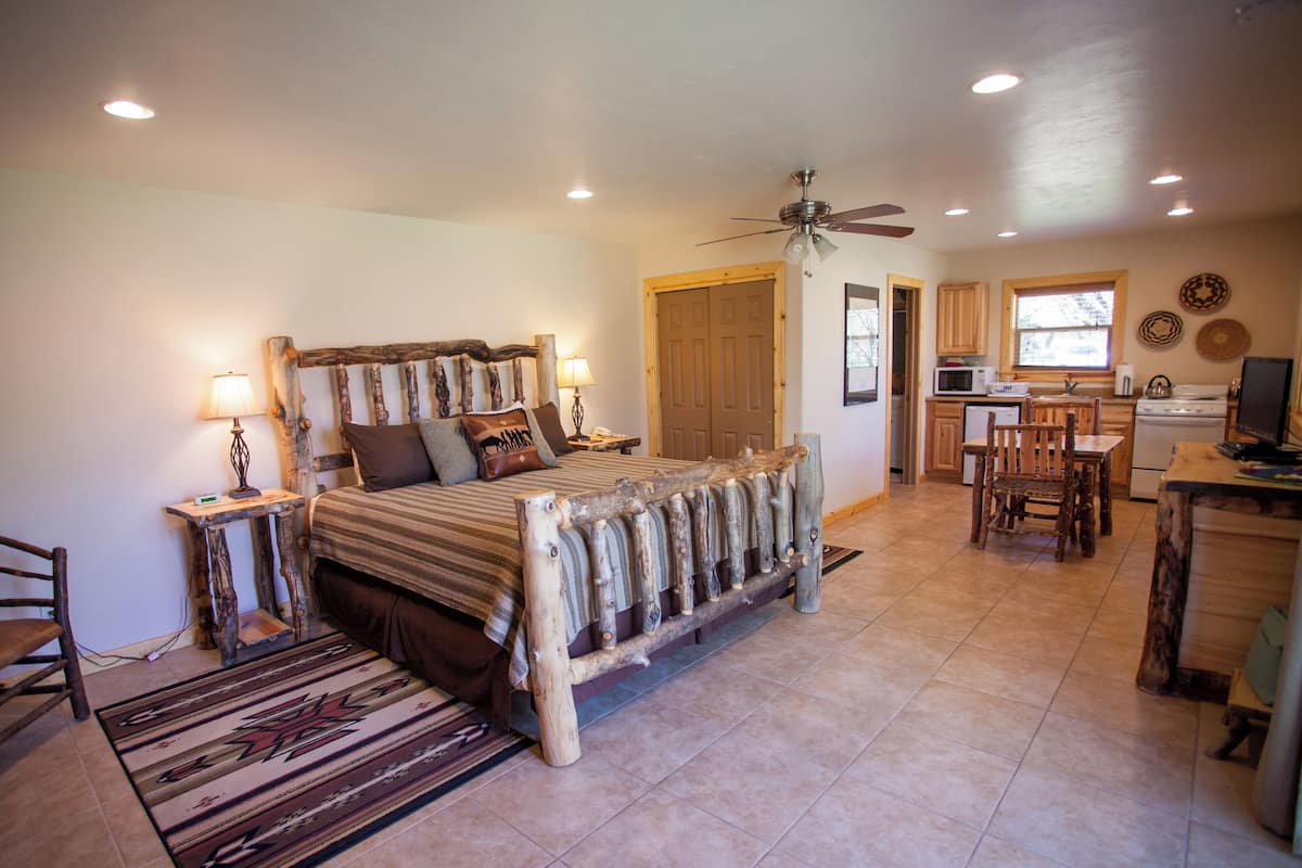 A bedroom with a bed, bedside tables, a wooden headboard, a ceiling fan, and a view into the adjacent kitchen.