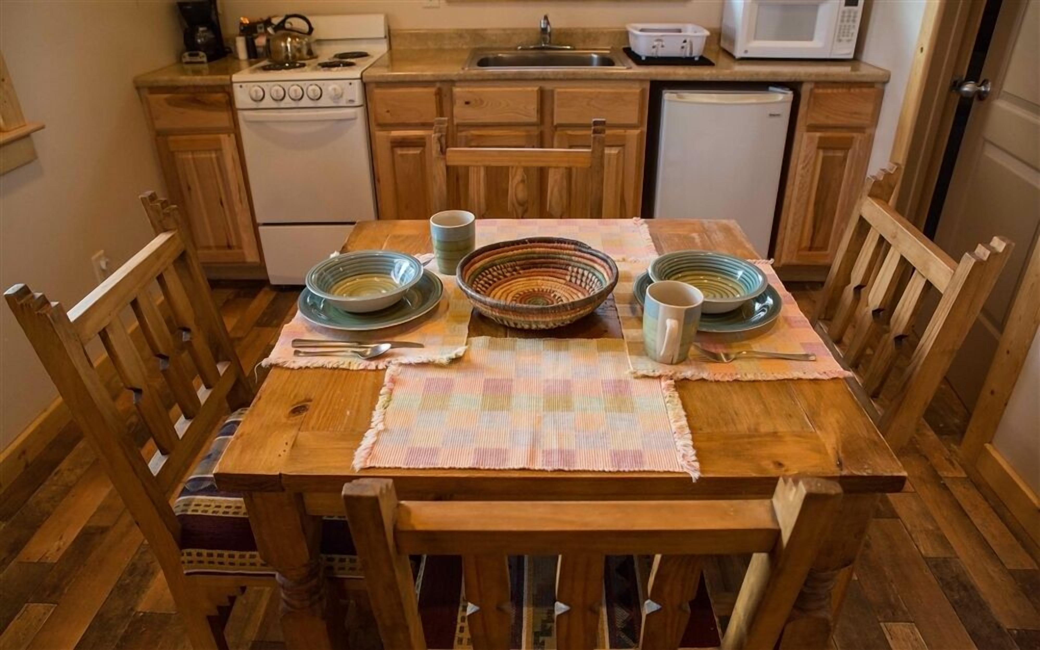 Una mesa de comedor de madera con platos, cuencos y cubiertos, en una cocina con armarios y electrodomésticos de madera.