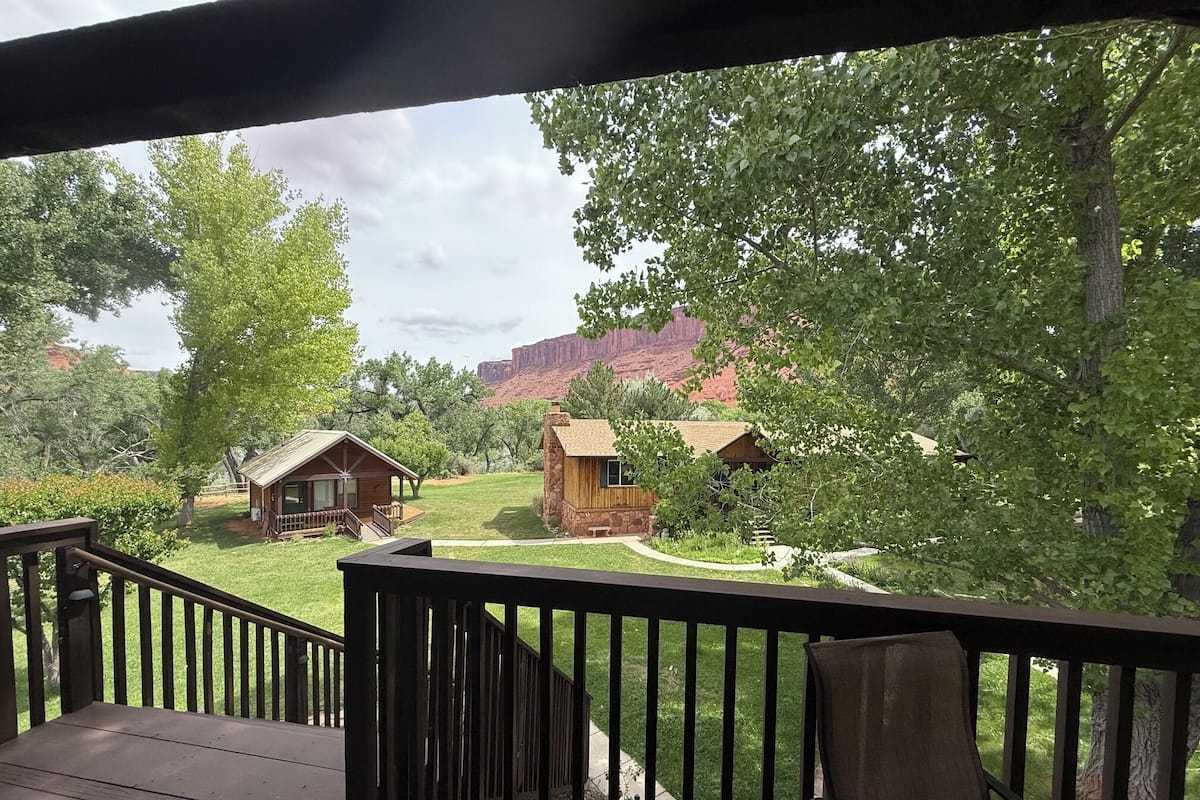 A wooden deck with a railing, a grassy lawn, and a view of rustic cabins in a wooded area.
