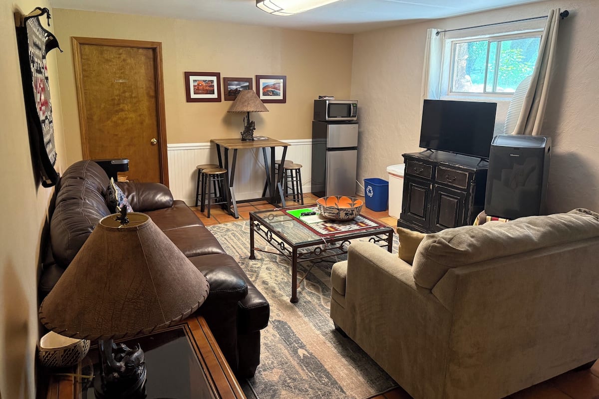 A living room with a brown leather couch, a beige armchair, a coffee table with decorative items, a TV, a refrigerator, and a window with curtains.
