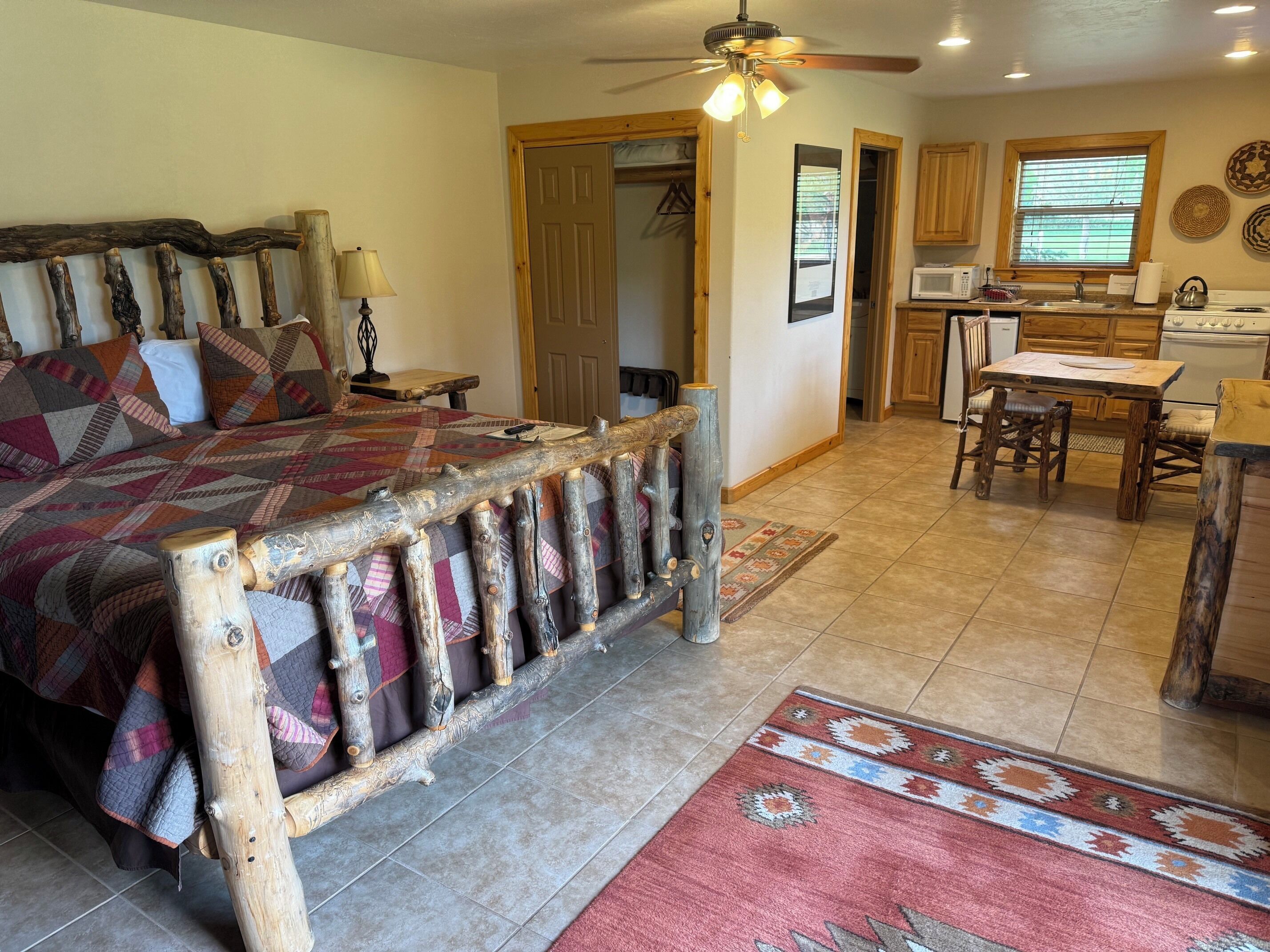 A bedroom with a wooden headboard, a bed with a patterned quilt, a nightstand, a lamp, and a ceiling fan. Adjacent is a kitchen with wooden cabinets, a microwave, a sink, and a dining area with a table and chairs.