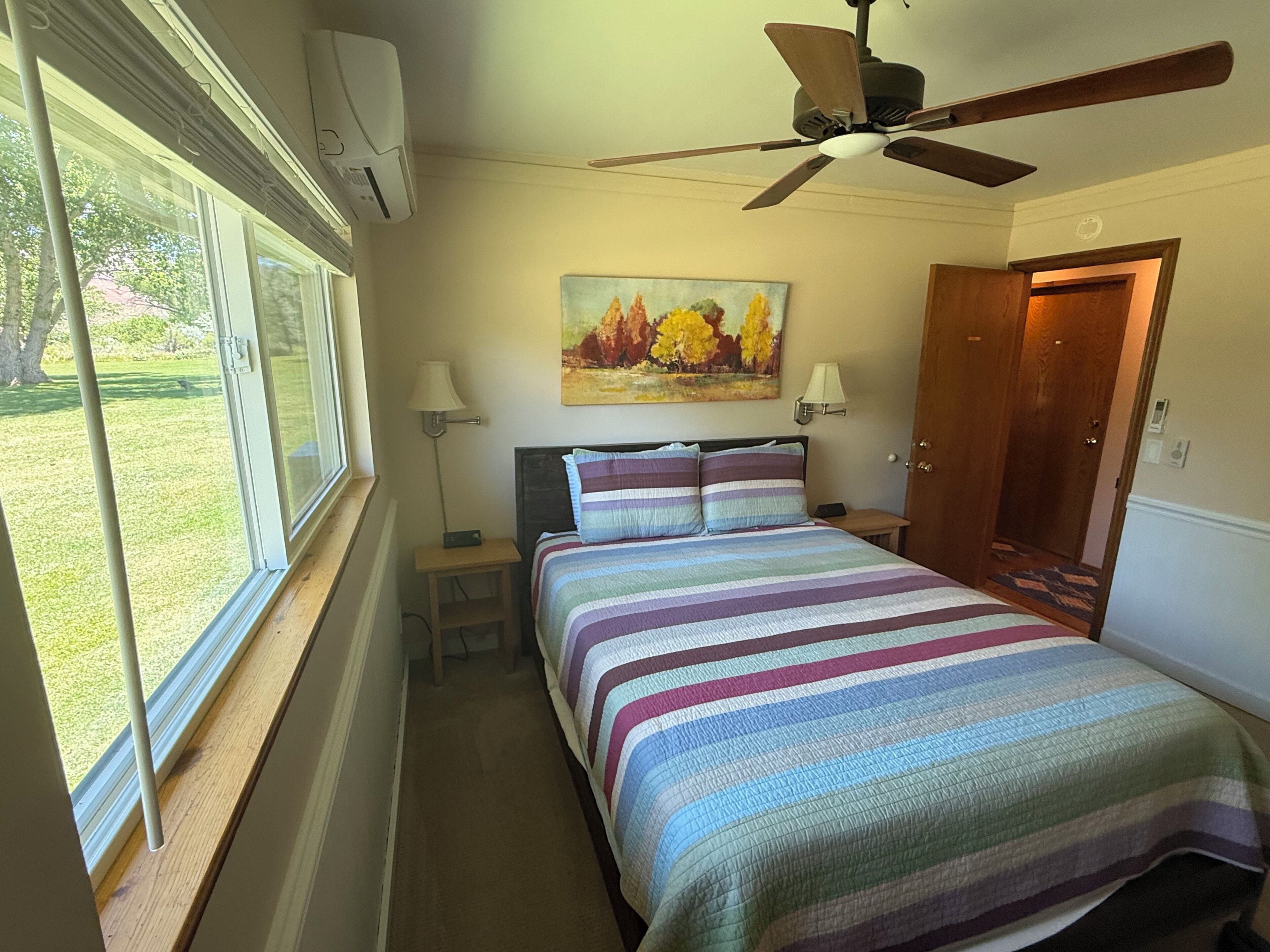 Ein Schlafzimmer mit einem Bett, einem Deckenventilator, einem Gemälde, einem Nachttisch, einer Lampe und einer Tür.