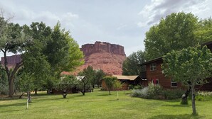A rustic cabin with a balcony, surrounded by trees and a grassy lawn, with a red rock formation in the background.
