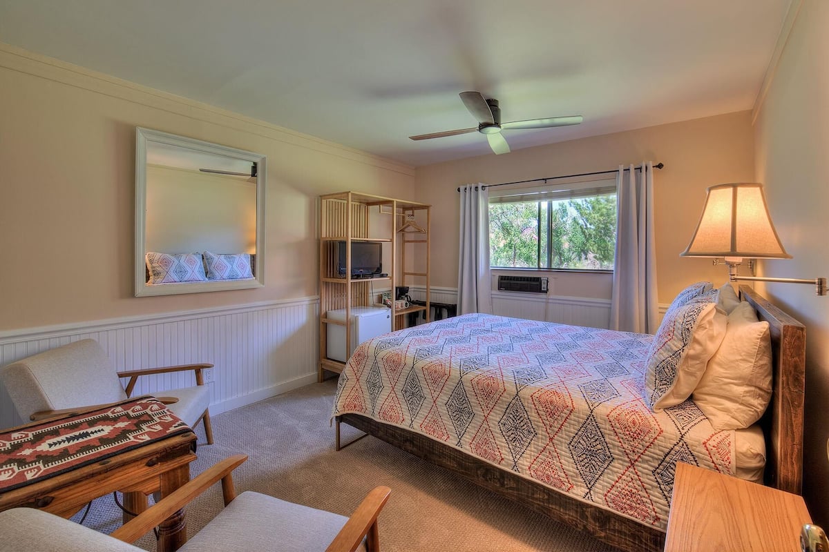 A bedroom with a bed, a chair, a desk, a mirror, a ceiling fan, a window with curtains, and a lamp.