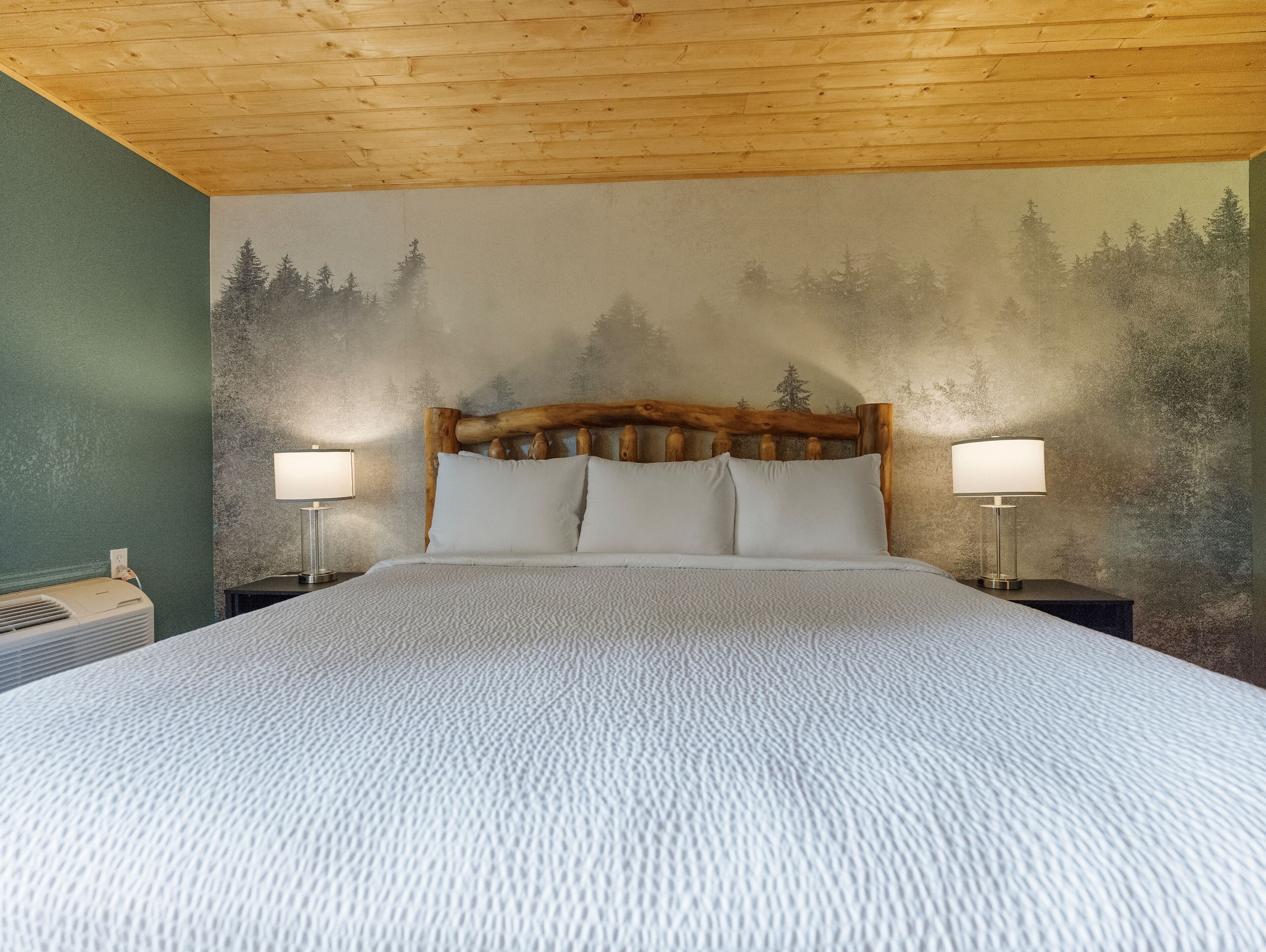 A bedroom with a large bed, wooden headboard, two bedside lamps, and a forest mural on the wall.