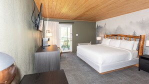 A hotel room with a wooden ceiling, a bed with white bedding, a desk with a lamp, and a view of a forest through a window. - Renovated King ADA Pet-Friendly room (Townsend)