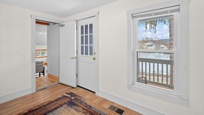A room with a wooden floor, white walls, a white door, and a window with a view of a balcony and trees. - The Barn - Lake View and Pet Friendly (Pittsfield)