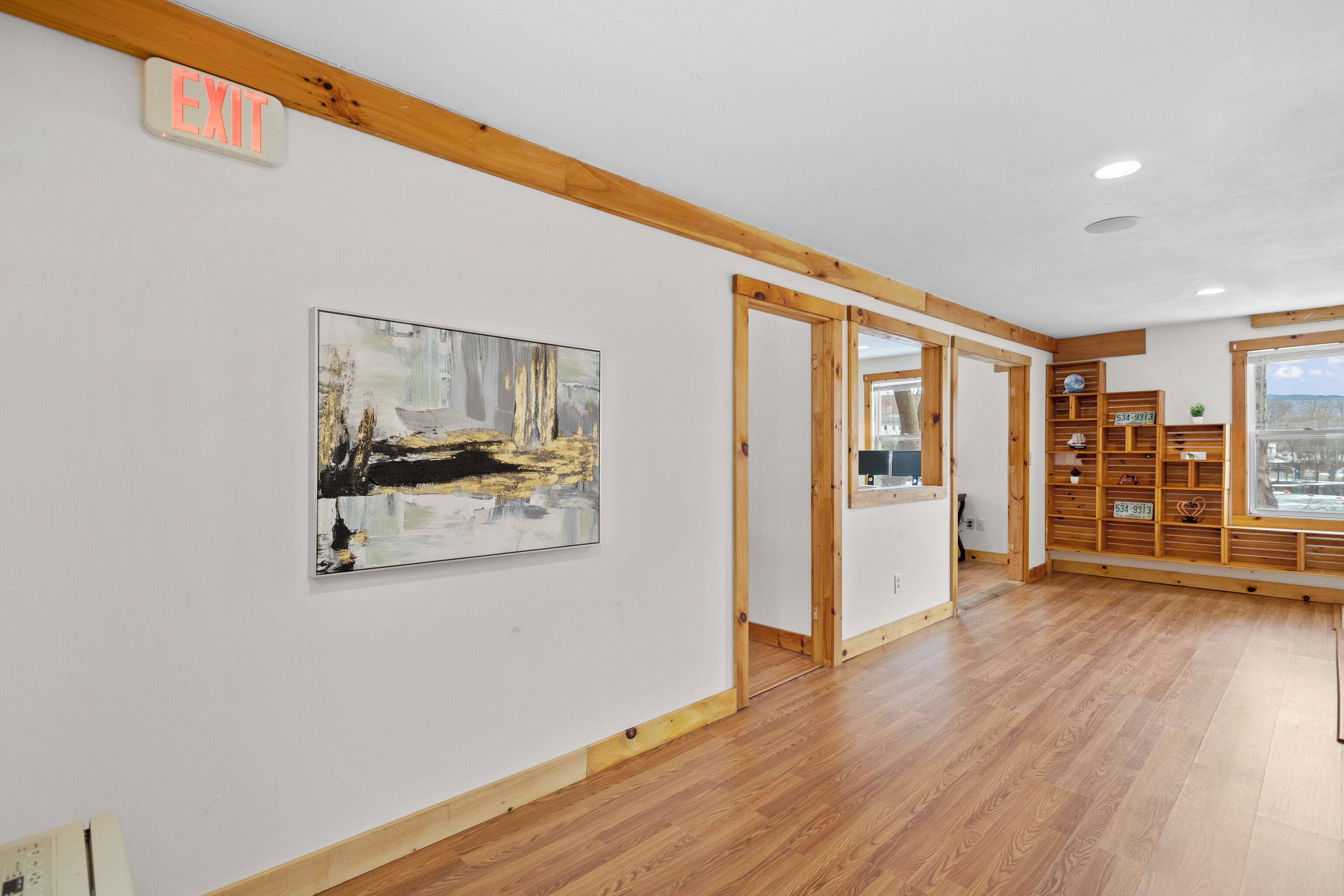 Une chambre d’hôtel avec un plancher en bois, un tableau au mur, une étagère en bois avec quelques objets, et un panneau indiquant la sortie.