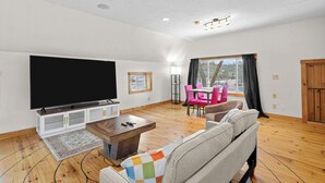 A living room with a flat-screen TV, wooden flooring, a sofa, a coffee table, and a dining area with pink chairs. - The Barn - Lake View and Pet Friendly (Pittsfield)